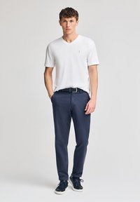 Vit bomull kortärmad V-ringad skjorta i kombination med marinblå chinos och svarta sneakers. Bältet är vävt med en silverfärgad spänne.