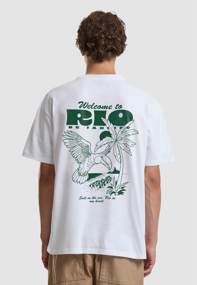 Person wearing white t-shirt with green print of toucan, palm tree, beach umbrellas, and text "Welcome to Rio de Janeiro" on back.