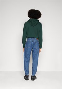 Grön huvtröja med stor huva, tillsammans med lösa blå jeans med bakfickor. Svarta skor fulländar outfiten. Enfärgad bakgrund.