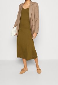 Olivově zelené, přiléhavé šaty spárované s béžovým blejzrem. Doplňky zahrnují bílou kabelku a hnědé ploché sandály. Hladká textilie.