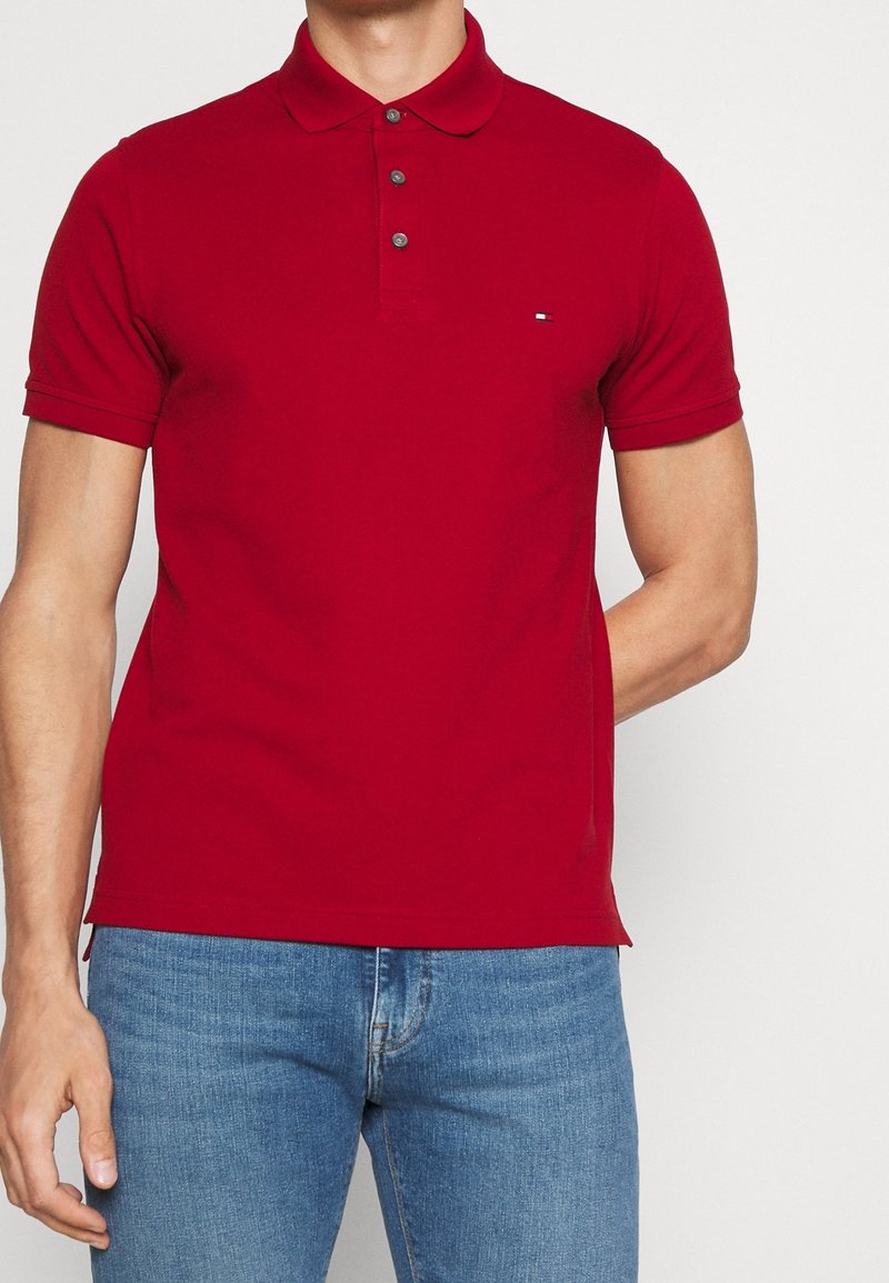 Homme portant un polo rouge ajusté avec trois boutons et un petit logo sur la poitrine, associé à un jean bleu en denim.