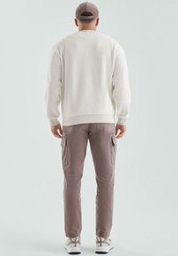 Mężczyzna w białym swetrze, taupe cargo pants, beżowych sneakersach i szarej czapce, stojący plecami na jednolitym tle.