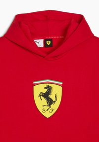 Rød hættetrøje med Ferrari-logoet, en sort oprejst hest på et gult skjold med "SF" og detaljer i grøn-hvid-rød.