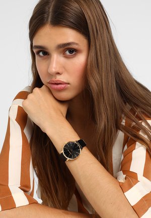 Black watch with a round face, gold accents, and a leather strap. Person with long hair shows the watch on their wrist, wearing a striped shirt.