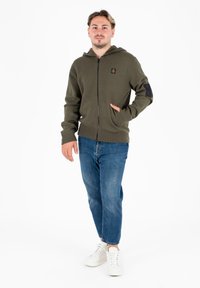 Felpa verde con zip, cappuccio con coulisse, polsini a coste e tasche laterali. Presenta una patch nera sul braccio sinistro e un logo sul petto.