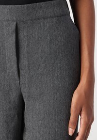 Szare spodnie w jodełkę z prostym przodem, wyposażone w pas i boczne kieszenie. Tkanina wydaje się mieć fakturę i strukturę. Ręka spoczywa na tkaninie.