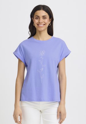 Woman smiling wearing a lavender t-shirt with a simple white line drawing of a rose and white pants against a plain background.