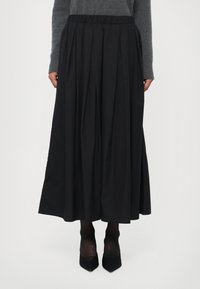 Svart, plisserad midi-kjol med elastisk midja, tillverkad av ett mjukt tyg. Bärs med genomskinliga svarta strumpbyxor och spetsiga klackar.