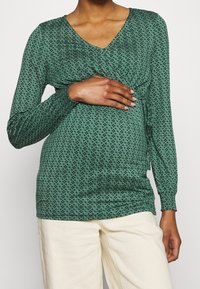 Zelený dlouhý rukáv top s výstřihem do V, se šperkovým vzorem a řasením v pase. Kombinováno s krémovými kalhotami.