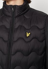 Czarna pikowana kamizelka z falistym wzorem szwów, zapinana na zamek, z podwyższonym kołnierzem i żółtym haftowanym logo orła na piersi.