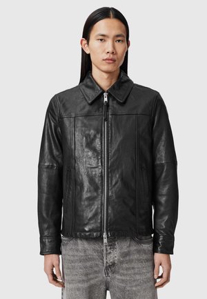 Młody mężczyzna z długimi ciemnymi włosami, ubrany w czarną skórzaną kurtkę na suwak oraz jasnoszare dżinsy, na jednolitym tle.