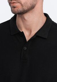 Musta polo-paita, jossa on teksturoitu kangas, kaksinappinen kaulus ja litteä kaulus; varustettu lyhyillä hihoilla ja hienovaraisella designilla.