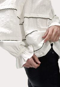Blusa blanca texturizada con ribete negro, con mangas abullonadas y detalle de puños fruncidos, combinada con pantalones negros ajustados.