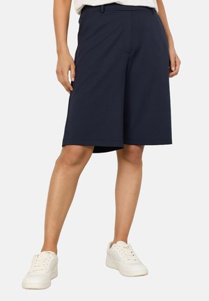 Marineblaue maßgeschneiderte Shorts aus einem glatten, dehnbarem Stoff, mit geradem Schnitt und Seitentaschen, kombiniert mit weißen Sneakern.