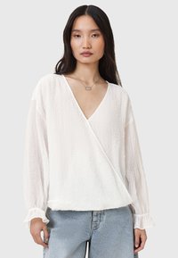 Hvid tekstureret bluse med lange ærmer og flæser ved ærmekanterne, parret med lyse denim. Har en dyb V-udskæring og en afslappet pasform.