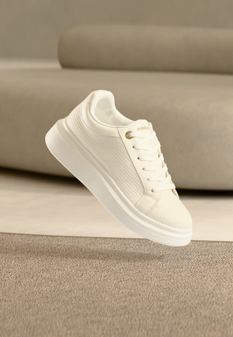 Λευκό χαμηλοκομμένο αθλητικό παπούτσι με υφασμάτινα πλαϊνά πάνελ, κορδόνια και παχιά λευκή σόλα, τοποθετημένο πάνω σε μπεζ χαλί και φόντο καναπέ.