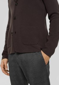 Dunkelbrauner Cardigan aus strukturiertem Stoff mit abgerundetem Saum, zwei vorderen Taschen und schwarzem Knopfverschluss. Kombiniert mit gemusterten grauen Hosen.