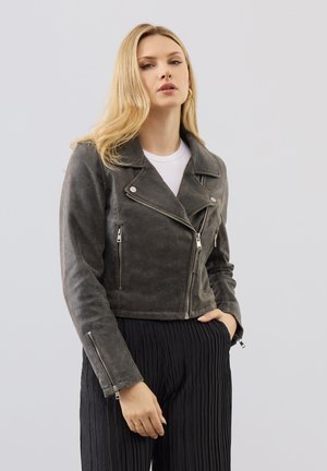 Mujer rubia que lleva chaqueta de motorista de ante gris oscuro con cremalleras sobre una camiseta blanca y pantalones negros plisados, de pie contra un fondo liso.