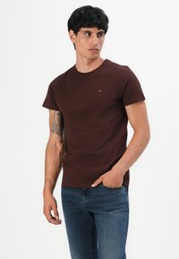Mann mit dunklen Haaren, der ein braunes T-Shirt mit kurzen Ärmeln und blaue Jeans trägt. Er steht mit einer Hand in der Tasche und sieht leicht zur Seite.