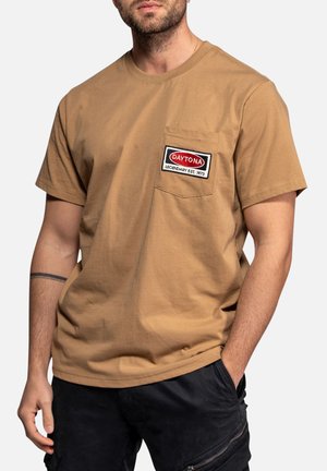 Homme portant un T-shirt marron à manches courtes avec une poche avant sur laquelle est écrit "Daytona Legendary Est. 1973" et un pantalon noir avec une poche zippée.