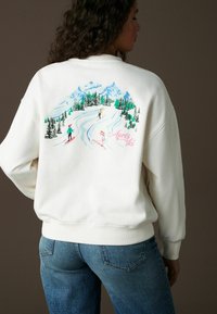 Kermanhuppari, jossa on värikäs hiihtokohtaus takana, mukana vuoria, puita ja hiihtäjiä. Ribbaatut hihansuut ja helma antavat tekstuuria.