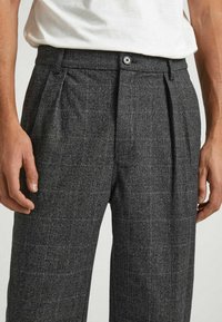 Grå mönstrade byxor med veckad front, tillverkade av ett texturerat tyg. Har en knappstängning och sidofickor.
