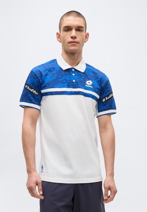 Νέος άνδρας με λευκό και μπλε πόλο μπλουζάκι Lotto με γεωμετρικά σχέδια, στέκεται μπροστά σε απλό φωτεινό φόντο.