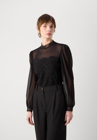 Zwarte doorzichtige blouse met kantdetails op het lijf en de manchetten, hoge hals en puffed lange mouwen. Gecombineerd met hoogzitende zwarte broeken.