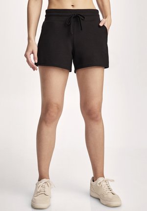 Woman wearing black drawstring shorts and beige sneakers, standing with one hand in pocket against plain background.