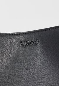 Zwart leren materiaal met een textuur oppervlak. Bevat het geperste "HUGO" logo. Afgeronde bovenkant met zichtbare stikdetails.