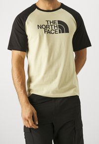 Kortærmet T-shirt med en lys beige krop og sorte raglanærmer, en rund halsudskæring og et fremtrædende sort logo på forsiden.