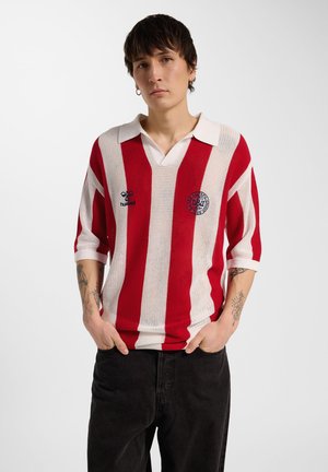 Osoba z krótkimi ciemnymi włosami ubrana w czerwono-biały pasiasty sweter z kołnierzykiem i czarne spodnie, stoi z rękami w kieszeniach.