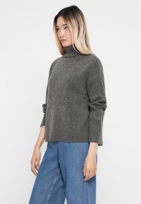 Pulover gri cu guler înalt dintr-un material tricotat moale, având o croială lejeră și un design ușor scurt. Asortat cu blugi evazați de culoare albastru deschis.