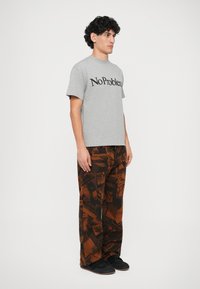 Maglietta in cotone grigia con la scritta "No Problem" stampata in nero in grassetto. Pantaloni marroni a fantasia con vari grafismi, indossati con sneakers nere.