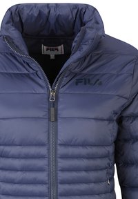 Päťkrát modrá prešívaná bunda s vysokým golierom, zapínaním na zips a logom Fila. Má horizontálny prešívaný dizajn a hladkú, lesklú textúru látky.