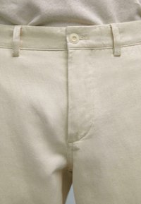 Beige cotton trousers with a smooth texture, featuring a button closure, belt loops, and a classic design. Close-up view of the waist area.