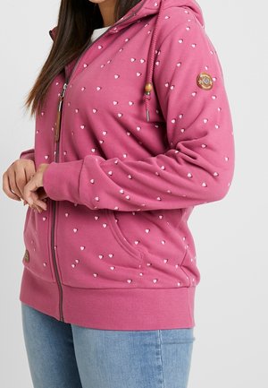 Mujer con sudadera rosa con cremallera con pequeños patrones de corazones blancos y vaqueros azules, manos juntas cerca de la cintura.