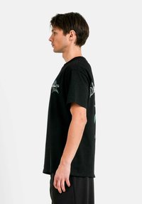 Svart bomullströja med korta ärmar, med grafisk design på både framtiden och baksidan, visad på en person i profil mot en enfärgad bakgrund.