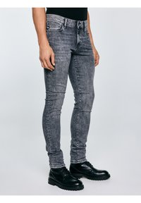 Isik, kes kannab tuhmunud halli kitsaid teksapükse ja musti nahast pitsidega saapaid, seisab helehallil taustal.