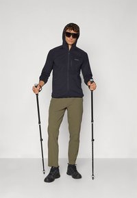 Zwarte capuchonvest met rits en capuchon, gecombineerd met lichtgroene broek en zwarte wandelschoenen; houdt wandelstokken vast; draagt een zonnebril.