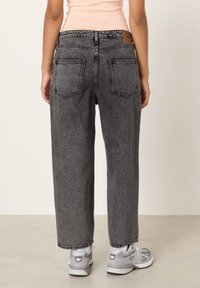 Sieviete tērpusies melnās, vidukļa augstumā augstās, brīva piegriezuma mazgātās džinsu biksēs ar ielāpa kabatām, valkā persiku krāsas topu un pelēku un baltu apavus, stāvot uz bēša grīdas.