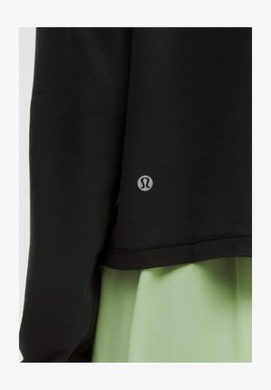 Zwart sporttop met een zachte textuur, horizontale zoom en een logo nabij de onderste rand. Gecombineerd met een lichtgroene broek.