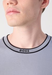 Grå T-shirt med en rund sort og lys grå ribbet krave og et logodetalje i halsen. Glat tekstur og tætsiddende design.