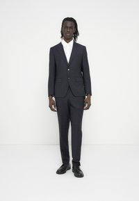 Tamsiai mėlynas kostiumas su prigludusiu švarku ir kelnėmis, pagamintas iš lygios medžiagos, dėvimas ant balto marškinių ir derinamas su juodais batais.