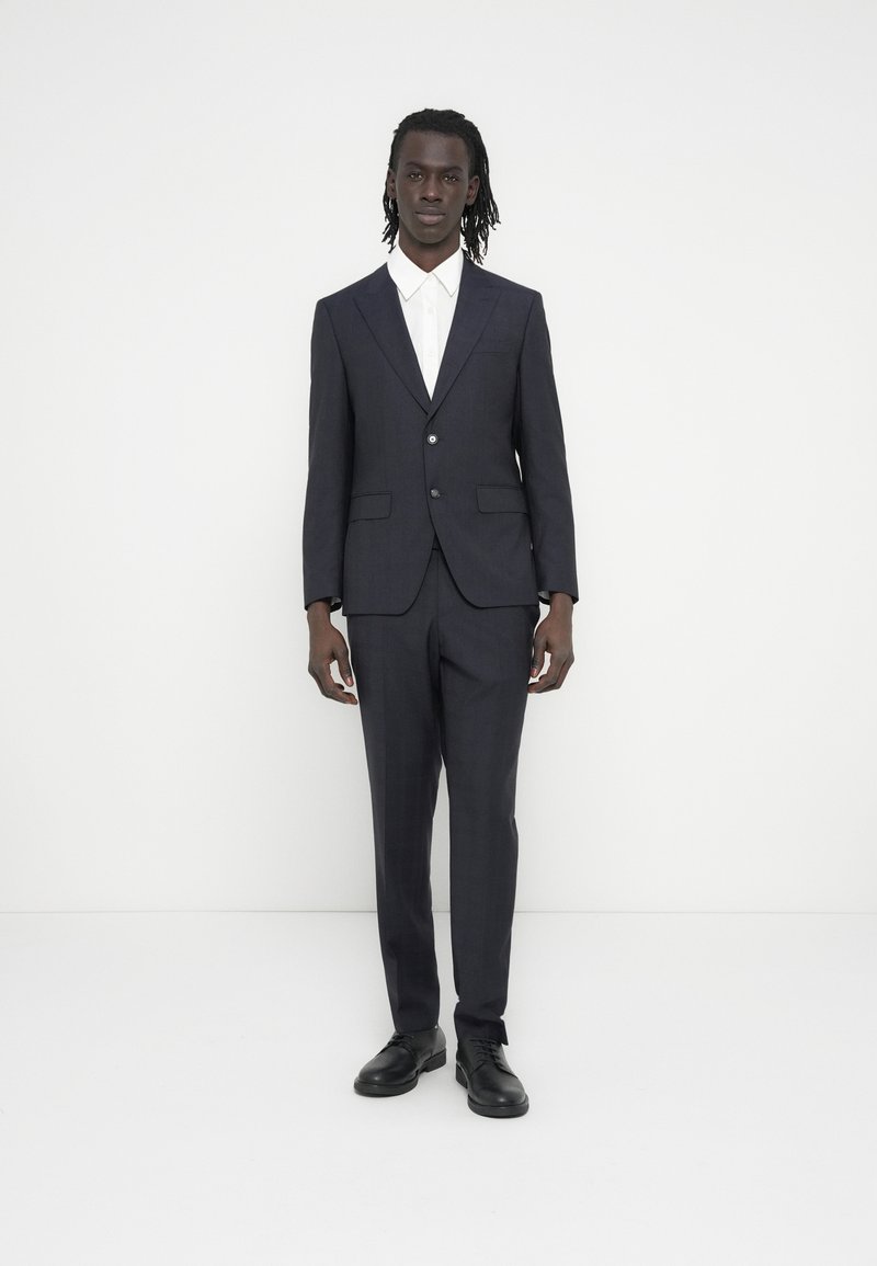 Tamsiai mėlynas kostiumas su prigludusiu švarku ir kelnėmis, pagamintas iš lygios medžiagos, dėvimas ant balto marškinių ir derinamas su juodais batais.