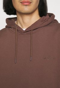 Brauner Baumwoll-Hoodie mit großzügiger Kapuze, Kordelzügen und gesticktem Logo auf der Brust. Glatte Textur und entspannter Schnitt.