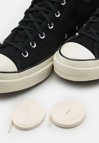 Svarta tygskor med vita gummikläppar, metallhål, och två rullar krämfärgade skosnören bredvid dem på en vit bakgrund.