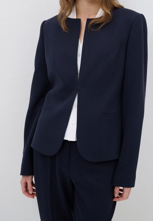 Kvinde iført en skræddersyet navy blazer over en hvid button-up skjorte og matchende navy bukser mod en ensfarvet lys baggrund.