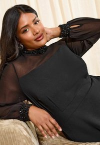 Rochie neagră cu un top transparent, împodobită cu accente de paiete la guler și manșete. Material neted cu o siluetă ajustată și buzunar.