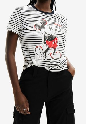 Femme portant un t-shirt rayé blanc et noir avec un motif Mickey Mouse rouge et noir, associé à un pantalon noir et une bague argentée à la main gauche.